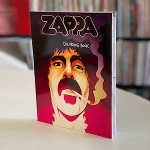 Puede incluir: Un libro para colorear de Zappa con un retrato de Frank Zappa en la portada, fumando un cigarrillo. La portada tiene la palabra "Zappa" en letras grandes y estilizadas sobre las palabras "Libro para colorear". El fondo es una mezcla de rojo y morado.