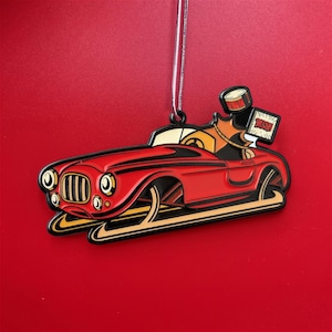 May include: A red car ornament with black accents and a gold-colored base. The ornament features a cartoon design of a car with a driver and a drum. The sign says "RUSH". The ornament hangs from a silver string against a red background.