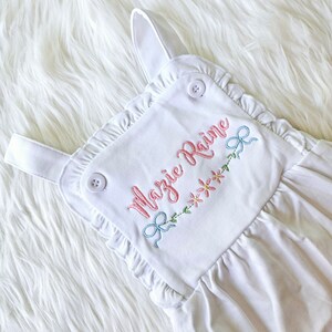 Pode incluir: Macac&atilde;o branco para beb&eacute; com acabamento em folhos e detalhes bordados. O macac&atilde;o apresenta o nome "Mazie Raine" em escrita rosa, juntamente com flores rosa e la&ccedil;os azuis. Dois bot&otilde;es brancos prendem as al&ccedil;as.