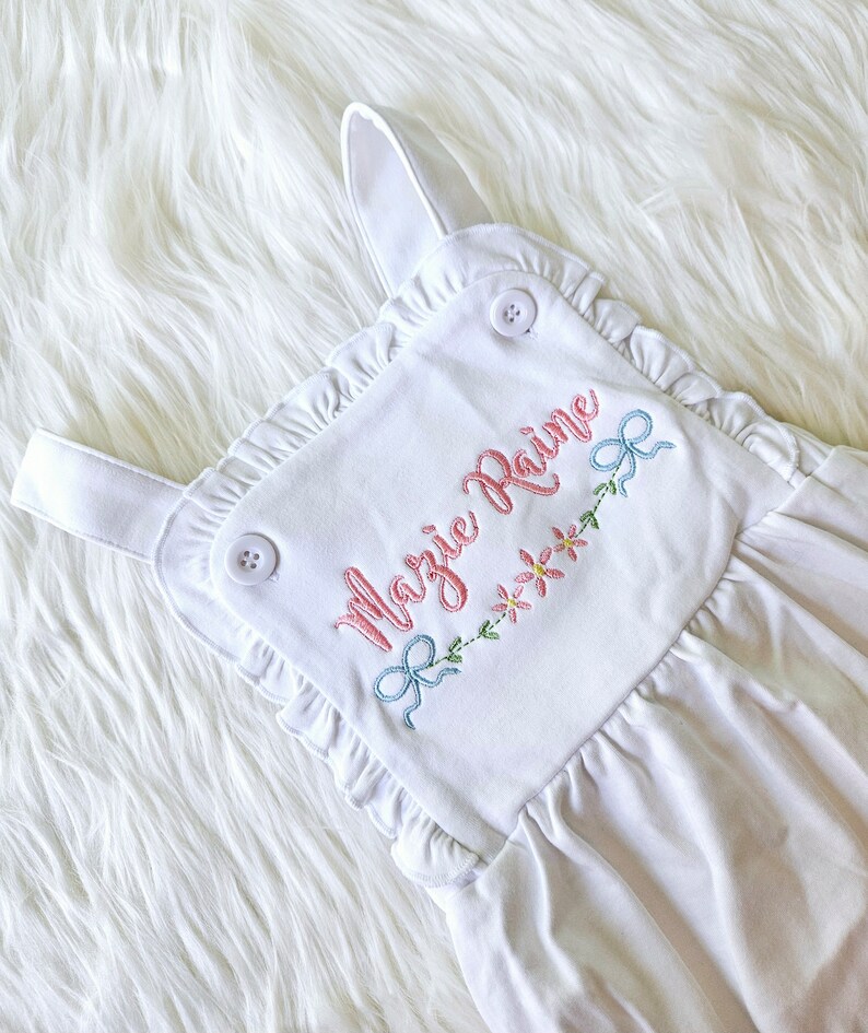 Pode incluir: Macac&atilde;o branco para beb&eacute; com acabamento em folhos e detalhes bordados. O macac&atilde;o apresenta o nome "Mazie Raine" em escrita rosa, juntamente com flores rosa e la&ccedil;os azuis. Dois bot&otilde;es brancos prendem as al&ccedil;as.