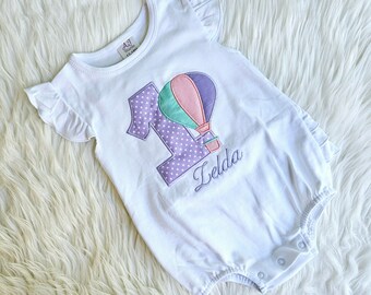 May include: White onesie with a purple number one and a hot air balloon design. The onesie has a ruffle trim around the sleeves and the neckline. The name "Zelda" is embroidered below the balloon.