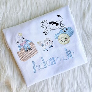 Puede incluir: Un pelele blanco para beb&eacute; con bordado azul de una vaca, dos personajes del libro infantil 'La oruga muy hambrienta', y un rey. El pelele est&aacute; personalizado con el nombre "Adam Jr."