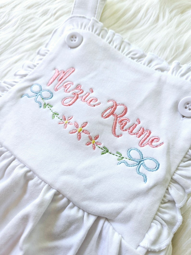 Pode incluir: Macac&atilde;o branco para beb&eacute; com bordas franzidas e al&ccedil;as. O macac&atilde;o apresenta o nome bordado "Mazie Raine" em rosa, juntamente com detalhes florais e la&ccedil;os em cores pastel. Dois bot&otilde;es brancos prendem as al&ccedil;as.