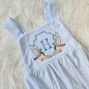 May include: Light blue baby overalls with white trim and embroidered ducks and the letter "H" in a crest.