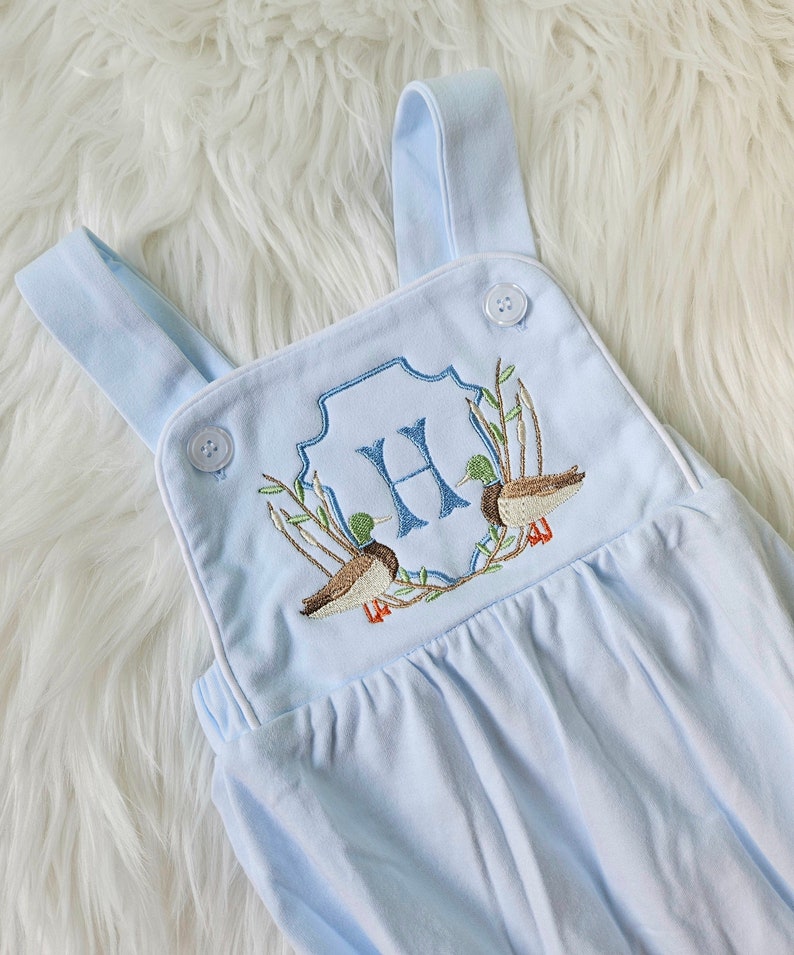 May include: Light blue baby overalls with white trim and embroidered ducks and the letter "H" in a crest.
