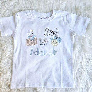Puede incluir: Camiseta blanca de manga corta con ilustraciones bordadas de una vaca, un hombre y un sol. El texto "Adam Jr" est&aacute; bordado debajo de las ilustraciones.