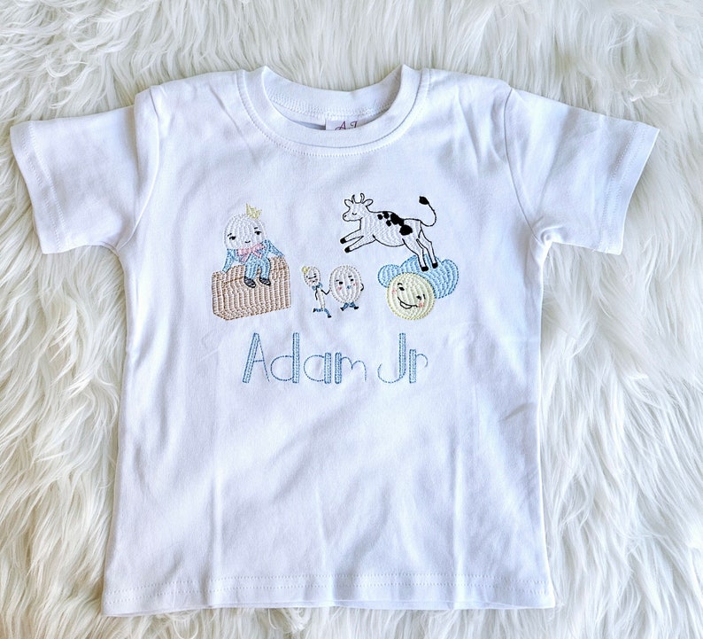 Puede incluir: Camiseta blanca de manga corta con ilustraciones bordadas de una vaca, un hombre y un sol. El texto "Adam Jr" est&aacute; bordado debajo de las ilustraciones.
