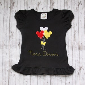 May include: Black t-shirt with a yellow, red, and white Mickey Mouse balloon design and the name "Nora Doreen" embroidered in yellow.
