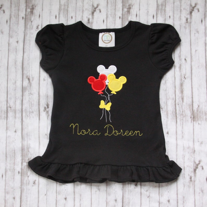 May include: Black t-shirt with a yellow, red, and white Mickey Mouse balloon design and the name "Nora Doreen" embroidered in yellow.