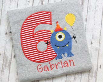 May include: A gray t-shirt with a red and white striped number six, a blue monster with one eye, a party hat, and a yellow balloon. The text "Gabrian" is embroidered below the monster.