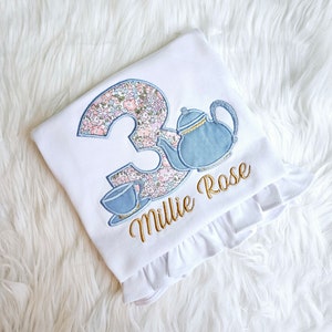 May include: White t-shirt with a floral number 3, a blue teapot, and a teacup. The name "Millie Rose" is embroidered in gold below the design.