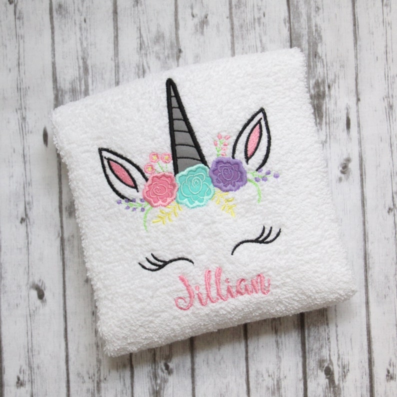 May include: White bath towel with an embroidered unicorn face with a floral crown and eyelashes. The name Jillian is embroidered below the unicorn.