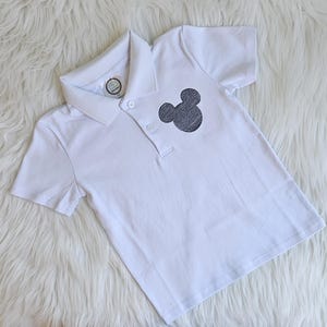 May include: White polo shirt with a gray Mickey Mouse silhouette on the chest. The shirt has a collared neckline and short sleeves. The shirt is laying on a white, fluffy surface.