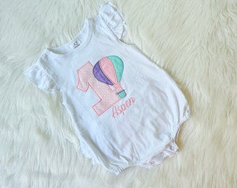 May include: White baby onesie with a pink and purple hot air balloon and the number one. The onesie has the name Aspen embroidered on it.