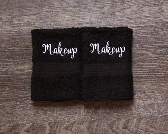 May include: Two black washcloths with the word "Makeup" embroidered in white script. The washcloths are made of a soft, textured material and are displayed on a wooden surface.