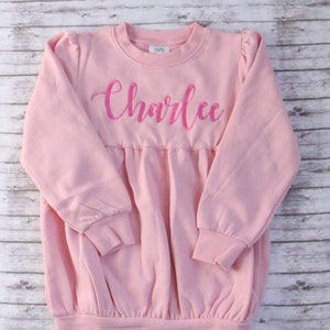 May include: A pink sweatshirt with a gathered waist and long sleeves. The sweatshirt has the name "Charlee" embroidered on the front in pink.