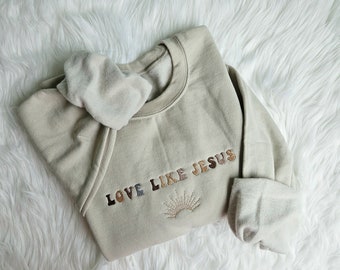 May include: A beige sweatshirt with the text "LOVE LIKE JESUS" embroidered in brown, black, and white letters. A small embroidered sun with rays is below the text.