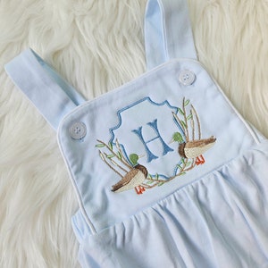 May include: Light blue baby overalls with white trim and embroidered ducks and a monogrammed "H" in a blue and green wreath.