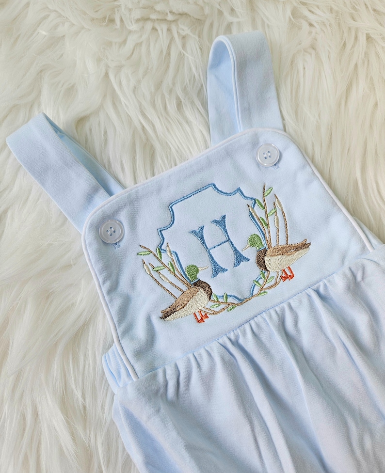 May include: Light blue baby overalls with white trim and embroidered ducks and a monogrammed "H" in a blue and green wreath.