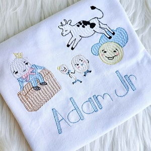 Puede incluir: Un pelele blanco con bordado azul de una vaca, dos huevos, un sol y un personaje del libro 'La oruga muy hambrienta'. El pelele tambi&eacute;n tiene el nombre 'Adam Jr' bordado en azul.
