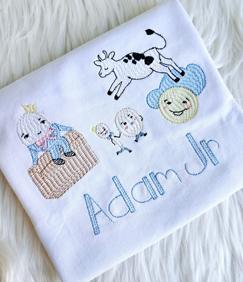 Puede incluir: Un pelele blanco con bordado azul de una vaca, dos huevos, un sol y un personaje del libro 'La oruga muy hambrienta'. El pelele tambi&eacute;n tiene el nombre 'Adam Jr' bordado en azul.