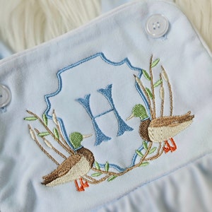 May include: Light blue baby onesie with an embroidered monogram of the letter "H" surrounded by two ducks and greenery.