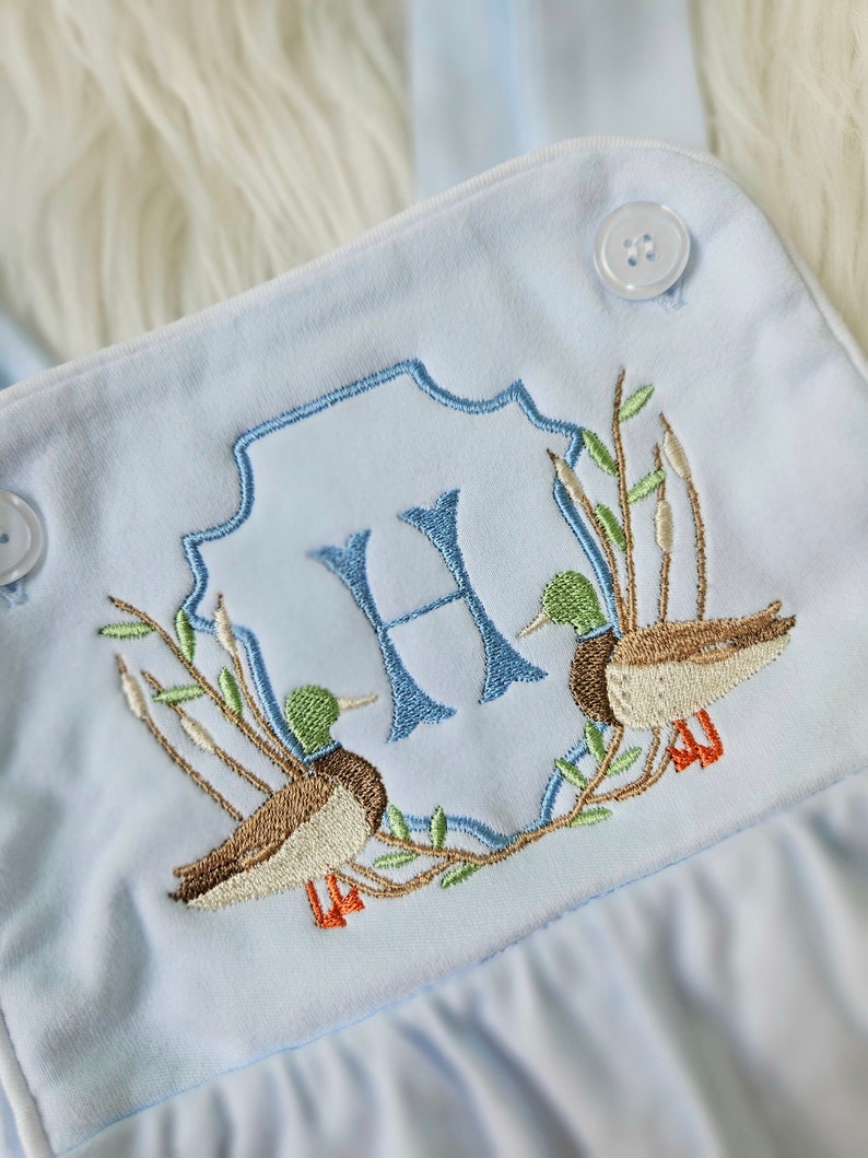 May include: Light blue baby onesie with an embroidered monogram of the letter "H" surrounded by two ducks and greenery.