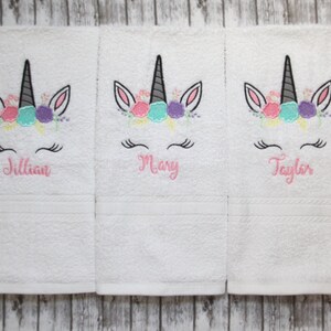 May include: Three white towels with embroidered unicorn faces and floral headbands. The towels are personalized with the names Jillian, Mary, and Taylor.