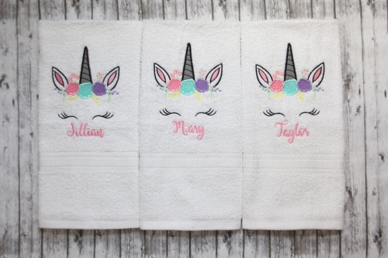 May include: Three white towels with embroidered unicorn faces and floral headbands. The towels are personalized with the names Jillian, Mary, and Taylor.