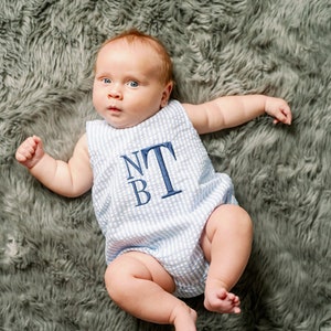 May include: A baby boy wearing a blue and white striped onesie with the letters "NTB" embroidered on the front. The baby is lying on a gray fuzzy blanket.