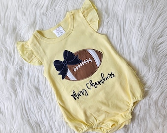 May include: Yellow baby onesie with a brown football and a black bow. The onesie has the text "Mary Chambers" embroidered on it.