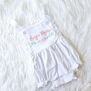 Pode incluir: Macac&atilde;o branco para beb&eacute; com acabamento em babados e detalhes de bot&otilde;es. O macac&atilde;o apresenta o nome bordado "Mazie Raine" em rosa, juntamente com detalhes florais e la&ccedil;os em cores pastel. A pe&ccedil;a tem al&ccedil;as ajust&aacute;veis e fechos de press&atilde;o.