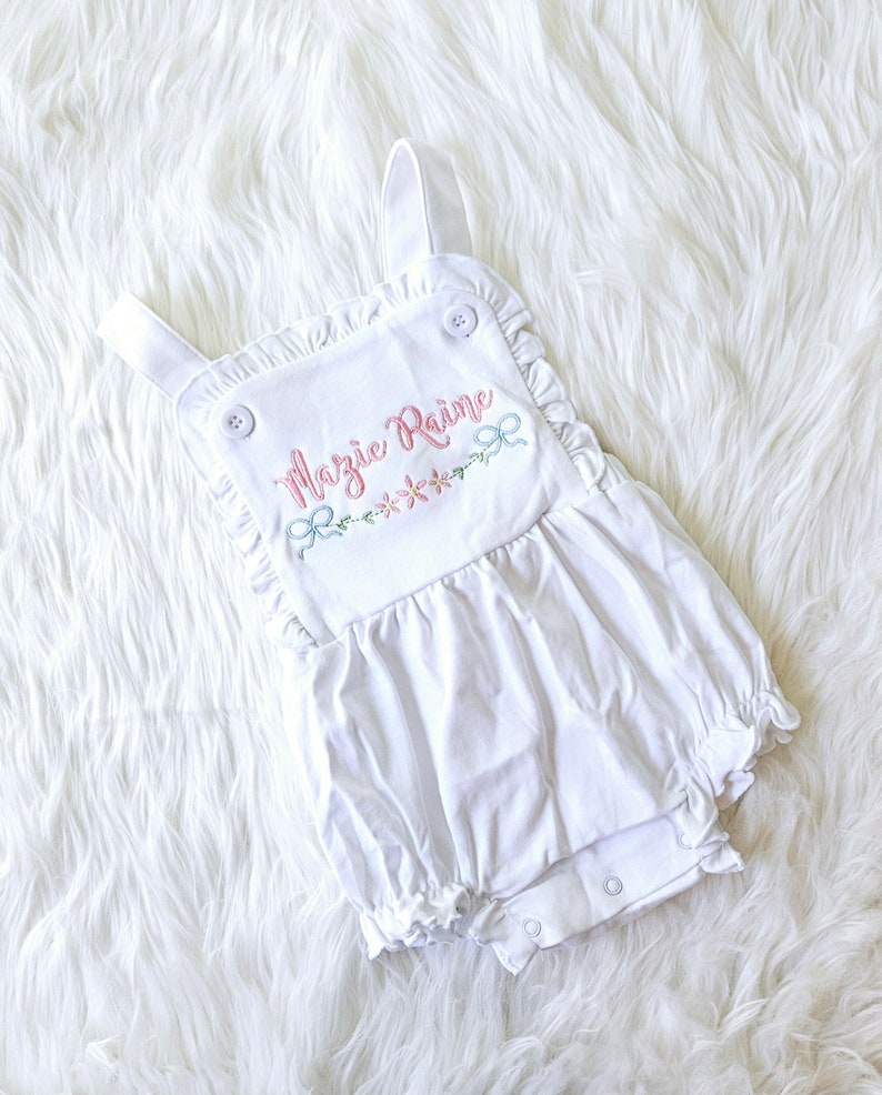 Pode incluir: Macac&atilde;o branco para beb&eacute; com acabamento em babados e detalhes de bot&otilde;es. O macac&atilde;o apresenta o nome bordado "Mazie Raine" em rosa, juntamente com detalhes florais e la&ccedil;os em cores pastel. A pe&ccedil;a tem al&ccedil;as ajust&aacute;veis e fechos de press&atilde;o.