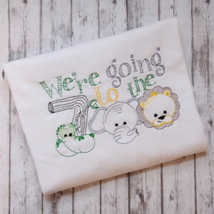 May include: White t-shirt with green, yellow, and black embroidery. The text "We're going to the Zoo" is embroidered on the shirt. There are also embroidered images of a lion, elephant, and frog.