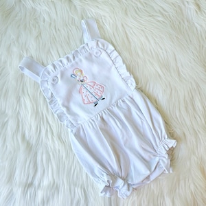 May include: White baby onesie with a ruffle trim and embroidered image of a girl holding a shepherd's crook.