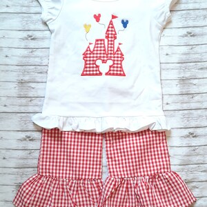 May include: A white short-sleeved shirt with a red and white gingham castle design and Mickey Mouse ears. The shirt has a ruffled hem. The shirt is paired with red and white gingham shorts with a ruffled hem.