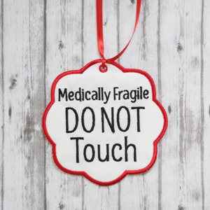 May include: A white, cloud-shaped tag with a red border and a red ribbon for hanging. The tag reads "Medically Fragile DO NOT Touch" in black text. The background is a light wood grain.