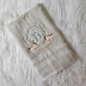 May include: A beige towel with an embroidered design featuring a blue letter "D" in a crest with two ducks and green leaves.