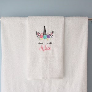 May include: White bath towel with a unicorn embroidered on it. The unicorn has a pink flower crown and the name "Alice" is embroidered below it.
