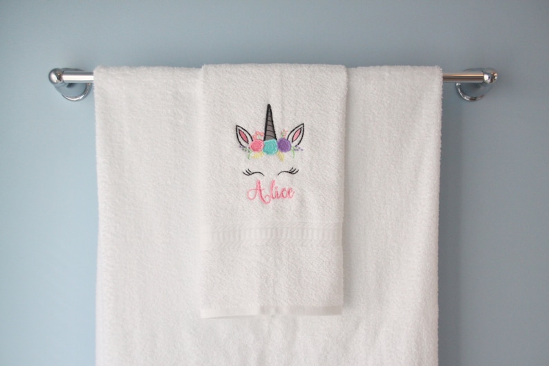 May include: White bath towel with a unicorn embroidered on it. The unicorn has a pink flower crown and the name "Alice" is embroidered below it.