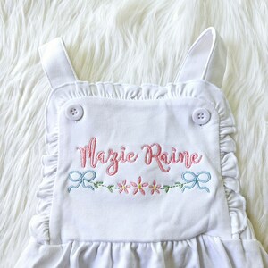 Pode incluir: Macac&atilde;o branco para beb&eacute; com acabamento em folhos e texto bordado "Mazie Raine" em rosa. O macac&atilde;o apresenta um design floral e de la&ccedil;o em rosa e azul, e &eacute; preso com dois bot&otilde;es brancos.