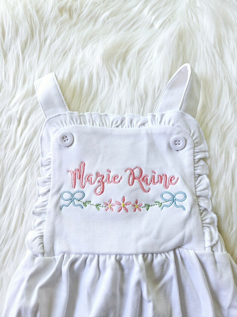 Pode incluir: Macac&atilde;o branco para beb&eacute; com acabamento em folhos e texto bordado "Mazie Raine" em rosa. O macac&atilde;o apresenta um design floral e de la&ccedil;o em rosa e azul, e &eacute; preso com dois bot&otilde;es brancos.