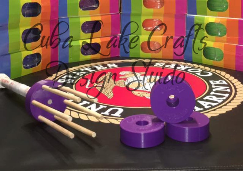 May include: Purple plastic bobbins with a hole in the center, stacked on top of each other. The bobbins are on a black surface with a red, white, and yellow design in the background.