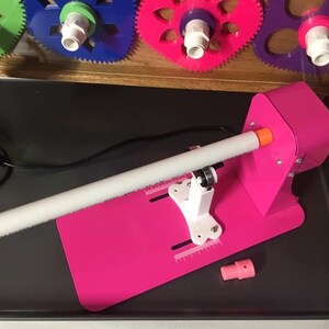 May include: A pink plastic machine with a white pipe and a white base. The machine has a black cord and a small pink nozzle. The machine is on a gray surface.
