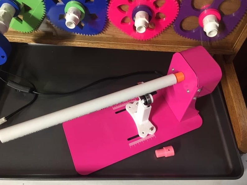 May include: A pink plastic machine with a white pipe and a white base. The machine has a black cord and a small pink nozzle. The machine is on a gray surface.