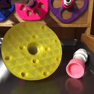 May include: A yellow plastic 3D printed round disc with a hole in the center and multiple smaller holes around the edge. A pink and white plastic connector is next to the disc.