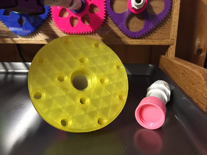 May include: A yellow plastic 3D printed round disc with a hole in the center and multiple smaller holes around the edge. A pink and white plastic connector is next to the disc.