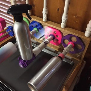 May include: A close-up of a DIY bottle drying rack with two silver spray bottles mounted on a wooden frame. The bottles are held in place by colorful plastic gears.