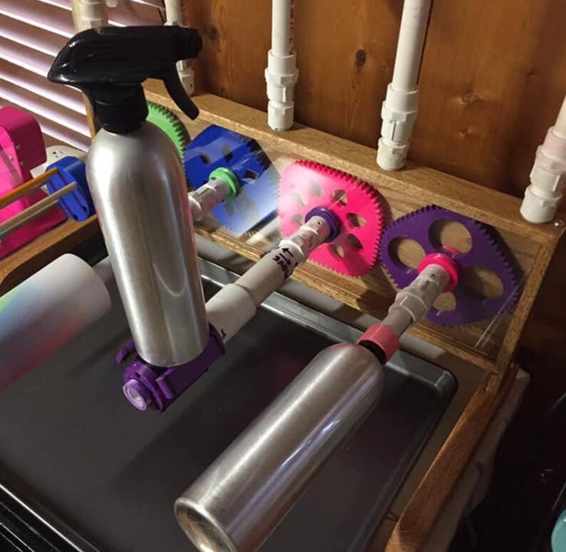 May include: A close-up of a DIY bottle drying rack with two silver spray bottles mounted on a wooden frame. The bottles are held in place by colorful plastic gears.