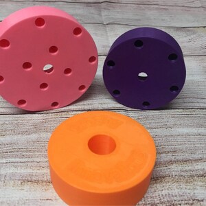 May include: Three round, colorful, plastic discs with different designs. The largest disc is pink with multiple holes, the middle disc is purple with a few holes, and the smallest disc is orange with a large hole in the center.